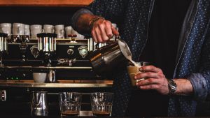 A barista pours coffee from a silver pitcher into a cup