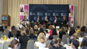 People on stage during the symposium, with the audience at tables in the foreground.