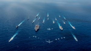 An aircraft carrier and a helicopter destroyer sail in formation with 16 other ships from the U.S. Navy and Japan Maritime Self-Defense Force as aircraft from the U.S. Air Force and Japan Air Self-Defense Force fly overhead in formation.