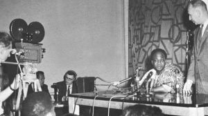 Kwame Nkrumah sits at a table and speaks into a microphone, while a camera films him.