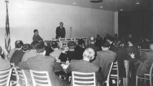 A man speaks to a room full of people in chairs.