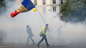 A person wearing a fluorescent yellow vest walks through smoke carrying a flag