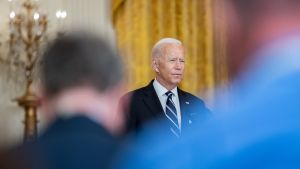 President Biden delivering remarks about COVID-19 response and vaccination program, blue suit partly blocking view in foreground.