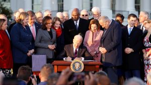 A crowd of White House officials surround President Joe Biden, watching as he signs the bill.