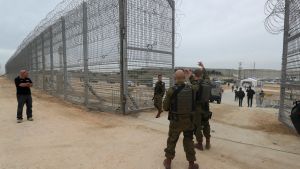 Guards stand near a large razor wire topped fence near Gaza on a cloudy day.