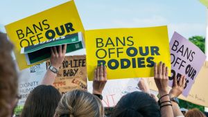 Hands hold yellow sign saying "Bans off our bodies" in front of a blue sky