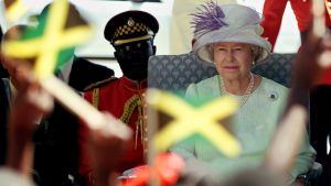 Queen Elizabeth II sits in a chair with people waving Jamaican flags in the foreground.