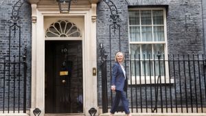 Liz truss walks outside from left to right in front of a brick building, door, and window.