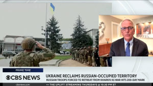 Screenshot of Ivo Daalder speaking on CBS news next to footage of Ukrainian soldiers saluting flag near pine trees.