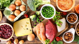 An assortment of food is laid out on a table, seen from a bird's eye view. Food includes eggs, cheese, peas, broccoli, and fish.