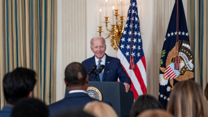 President Joe Biden delivers remarks at a podium before a crowd and in front of an American flag. 