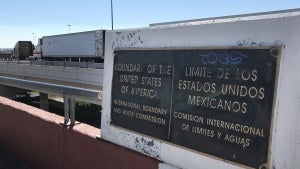 A sign marking the US-Mexico border, with semi trucks driving in the background