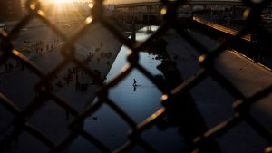 Asylum seeking migrants seen crossing water through a chain link fence at sunset.