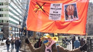 A man holds a "PUTIN IS A DICTATOR AND WAR CRIMINAL" sign attached to USSR flag