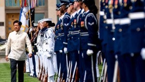 Marcos walks next to line of troops on green lawn.