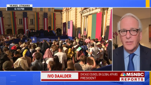 Screenshot of Ivo speaking on MSNBC at right, live footage of the empty stage in Vilnius with a crowd in front at left.