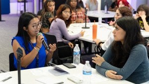 Student in blue shirt holds mic and gestures, seated at a table.