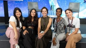 Lauren and team sit cross legged on edge of the stage and smile at camera.