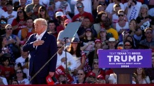 Donald Trump places hand over heart in front of a crowd in Waco, TX, podium with a sign bearing his name at right.