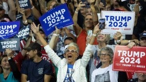 Crowd cheers and holds up "Trump 2024" signs, woman in a white jacket in the foreground with arms raised. 