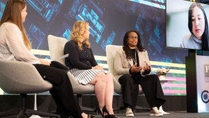 Three people sit in chairs while speaking on-stage, while someone joins virtually via TV screen.