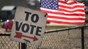 A sign reads "To Vote" above an arrow pointing right as an American flag flies in the background.