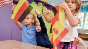 Small children at a classroom desk wave flags from the U.S., Germany, and Spain