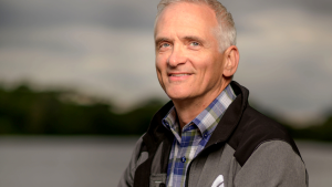 Headshot of a white man with gray hair, wearing a plaid shirt and gray fleece standing in front of a lake