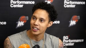 Brittney Griner sits in front of a black wall printed with WNBA logos speaking into a microphone