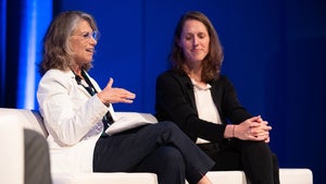 Two people sit in a white couch while talking on stage