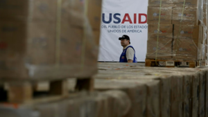 A person walks in front of a sign for USAID, with boxes in the foreground