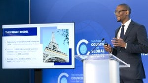 A man speaks at a podium with a TV screen beside him